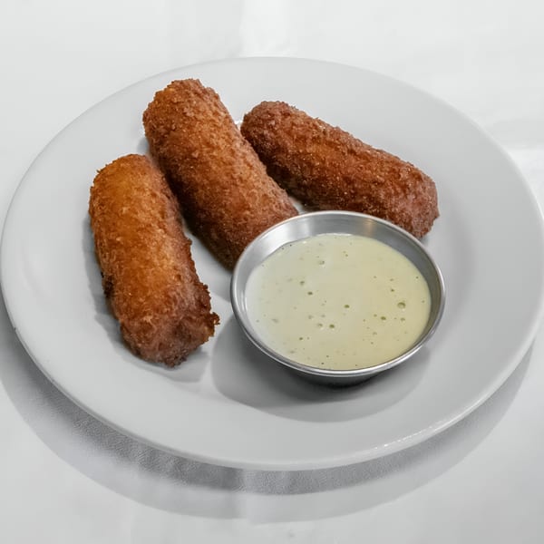 Three crispy Cuban croquettes with a side of creamy dipping sauce on a white plate.